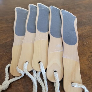 May include: Five wooden foot files with a light wood finish and dark gray abrasive surfaces. Each file has a foot-shaped design and a white rope loop for hanging. The files are arranged on a brown surface.