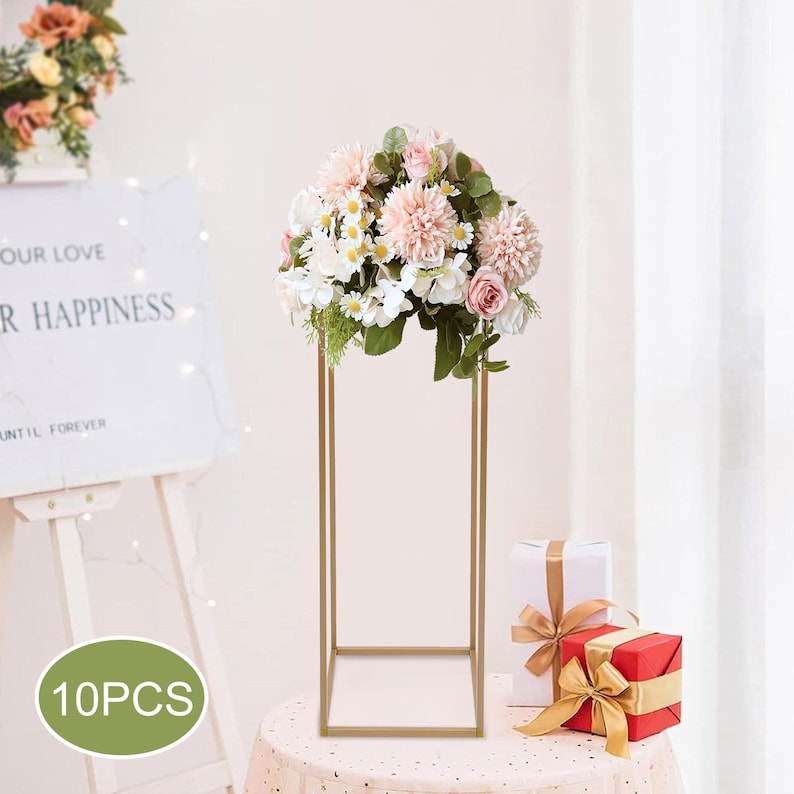 May include: A gold metal floral stand with a square base and a rectangular top, holding a bouquet of pink, white, and green flowers. The stand is on a round table with a pink tablecloth. Two wrapped gifts are on the table.