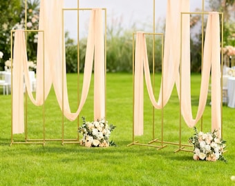 May include: Outdoor wedding decor featuring gold metal frames draped with peach fabric. The frames are arranged on a green lawn, with floral arrangements at the base. The scene is set in a garden, with a blurred background.