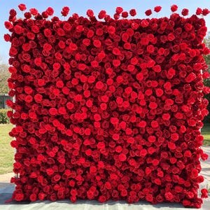 May include: A large square backdrop completely covered in vibrant red roses. The roses are densely packed, creating a textured surface. The backdrop is supported by a white frame, and the sky is visible in the background.