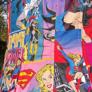 May include: A pink, blue, and red fabric corset with a patchwork design featuring various superhero characters, including Wonder Woman, Batgirl, and Supergirl. The fabric is quilted and has a pink stitching detail.
