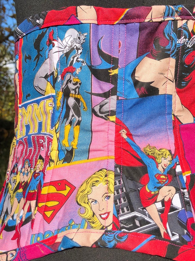 May include: A pink, blue, and red fabric corset with a patchwork design featuring various superhero characters, including Wonder Woman, Batgirl, and Supergirl. The fabric is quilted and has a pink stitching detail.