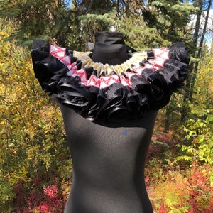 May include: A black and white ruffled collar with red and gold ribbon accents. The collar is made of fabric and is worn on a black mannequin.
