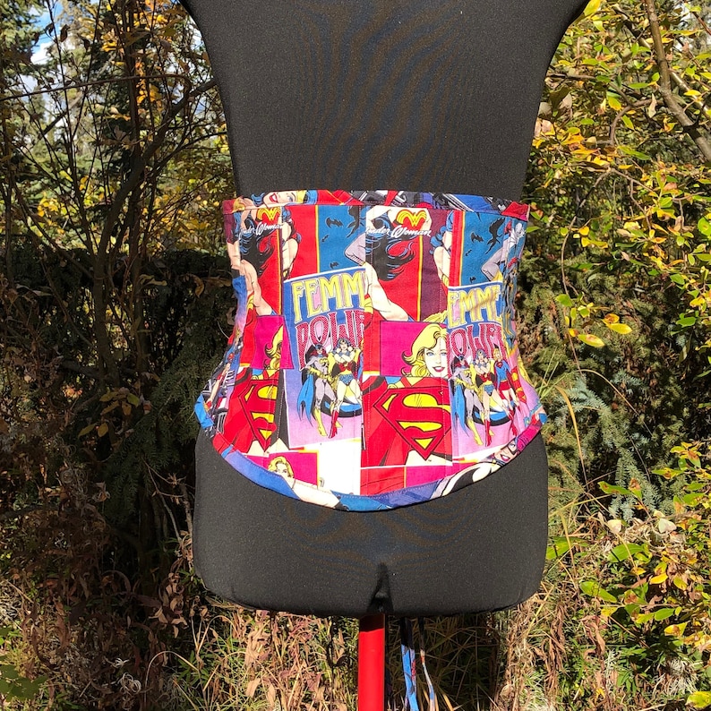 May include: A black corset with a colorful comic book print featuring female superheroes. The print includes the words "Femme Power" and images of Wonder Woman and Superman.