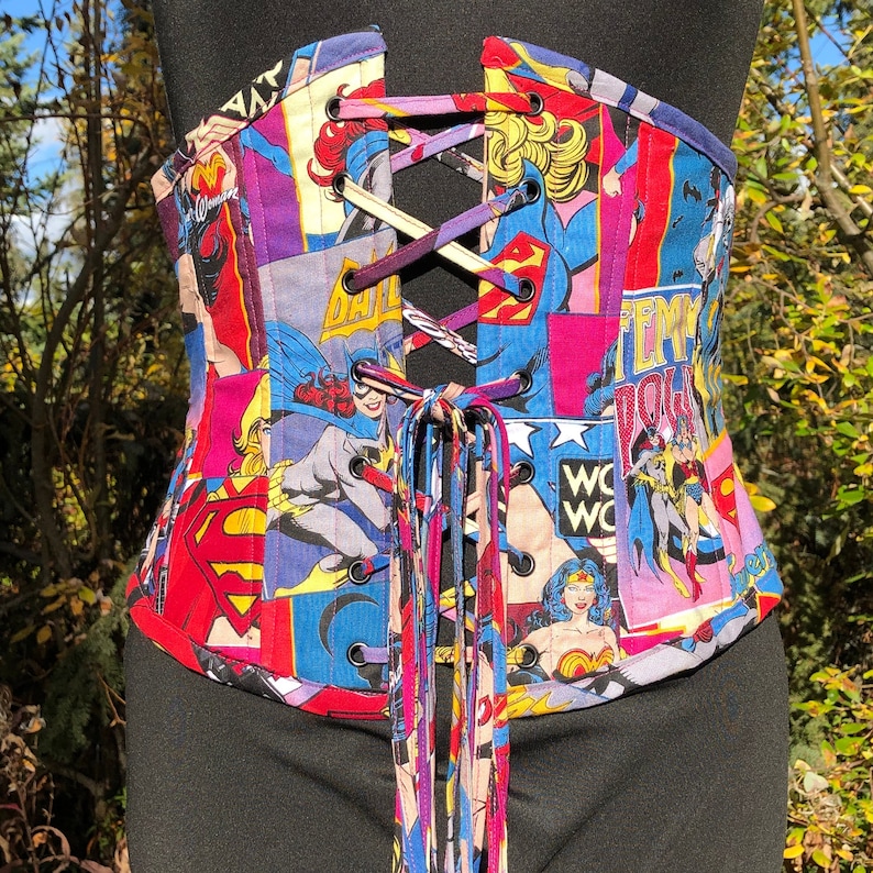 May include: A corset with a colorful print featuring various superhero characters, including Wonder Woman, Batgirl, and Supergirl. The corset is made of a fabric with a satin finish and has a lace-up closure in the front. The corset is decorated with a variety of colors, including red, blue, yellow, and green.