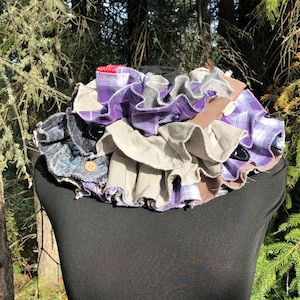 Puede incluir: Un collar de tela de varias capas con volantes en tonos de morado, gris y marrón. El collar está hecho de tela reutilizada y tiene un cierre de botón.