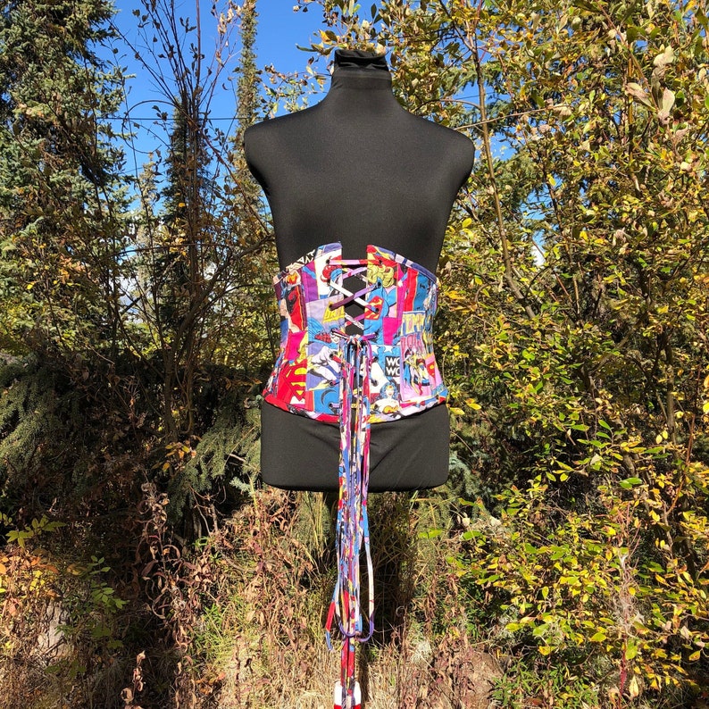 May include: A colorful corset with a comic book print. The corset is laced up the front and has long ties that hang down the back.
