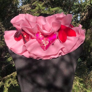 May include: A pink ruffled collar with three felt hearts attached. The hearts are red, pink with gold accents, and pink with sequins.
