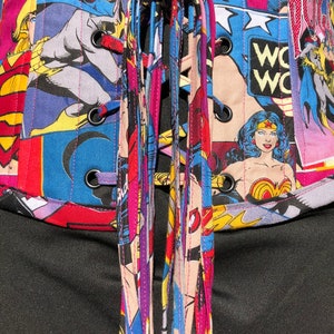 May include: A black corset with a colorful comic book print featuring Wonder Woman, Batgirl, and Supergirl. The corset has a lace-up back with multiple colorful laces.