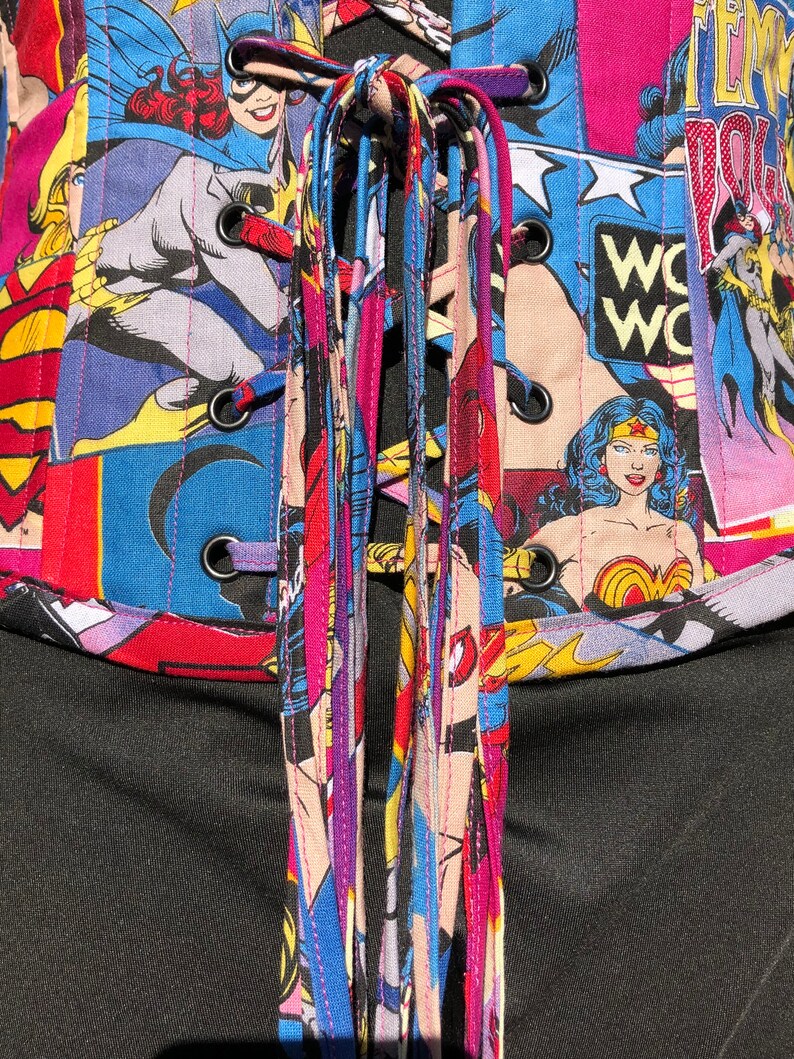 May include: A black corset with a colorful comic book print featuring Wonder Woman, Batgirl, and Supergirl. The corset has a lace-up back with multiple colorful laces.
