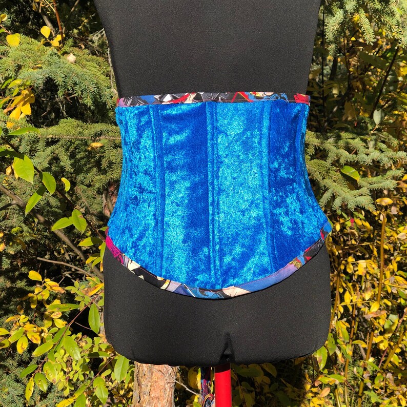 May include: A blue velvet corset with a colorful patterned trim. The corset is on a black mannequin.