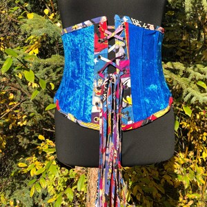 May include: A blue velvet corset with a colorful comic book print fabric trim. The corset has a lace-up front and a long tassel.