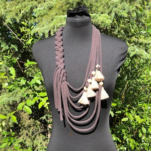 Könnte beinhalten: Eine braune Stoffkette mit mehreren Strängen und einem geflochtenen Abschnitt. Die Kette ist mit Holzperlen und beigefarbenen Quasten verziert.