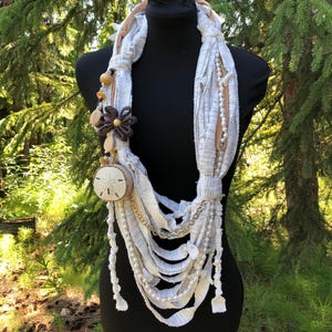 May include: A white, layered fabric necklace with wooden beads, a seashell, and a brown flower charm. The necklace is displayed on a black mannequin against a green background.