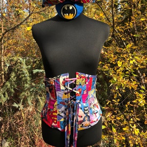 May include: A black corset with a colorful comic book print featuring superhero characters like Wonder Woman, Superman, and Batman. The corset has a lace-up front and a long red ribbon tie.