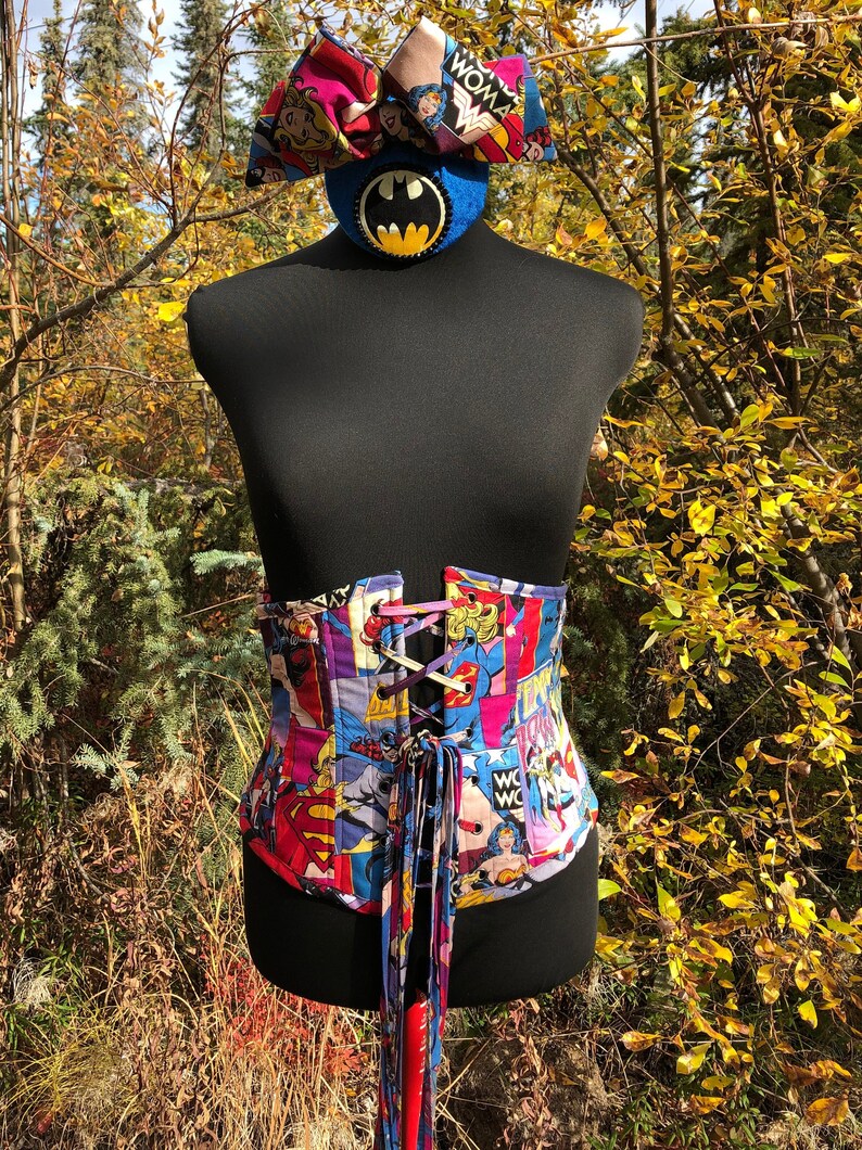 May include: A black corset with a colorful comic book print featuring superhero characters like Wonder Woman, Superman, and Batman. The corset has a lace-up front and a long red ribbon tie.