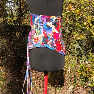 May include: A blue and pink corset with a comic book print featuring female superheroes. The corset has a long, fringed tie.