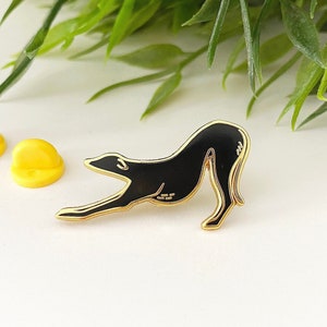 May include: Black enamel pin of a dog stretching, with a gold outline. The pin is on a white surface with two yellow rubber pin backs.