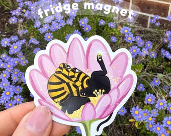 Pode incluir: Um íman de frigorífico com um gato preto com riscas amarelas, aninhado dentro de uma flor rosa. O íman tem a inscrição "fridge magnet". O fundo inclui flores azuis e uma parede de tijolos.