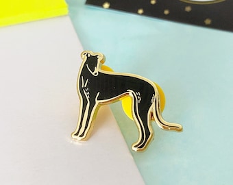 May include: A gold-colored enamel pin featuring a black Greyhound dog design. The pin has a yellow backing and is set against a light blue and white background. The pin is on a black card that says "Greyhound enamel pin".