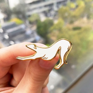 Pode incluir: Um broche de esmalte branco de um galgo em posi&ccedil;&atilde;o de corrida. O broche &eacute; dourado e tem um contorno detalhado.