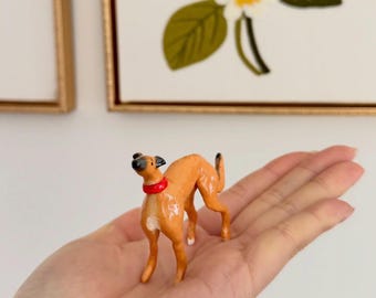 Pode incluir: Uma pequena estatueta pintada à mão de um cão castanho-claro com focinho preto e uma coleira vermelha brilhante. O cão está numa postura de alerta com a cabeça virada para cima, apresentando manchas brancas no peito e nas patas com acabamento.