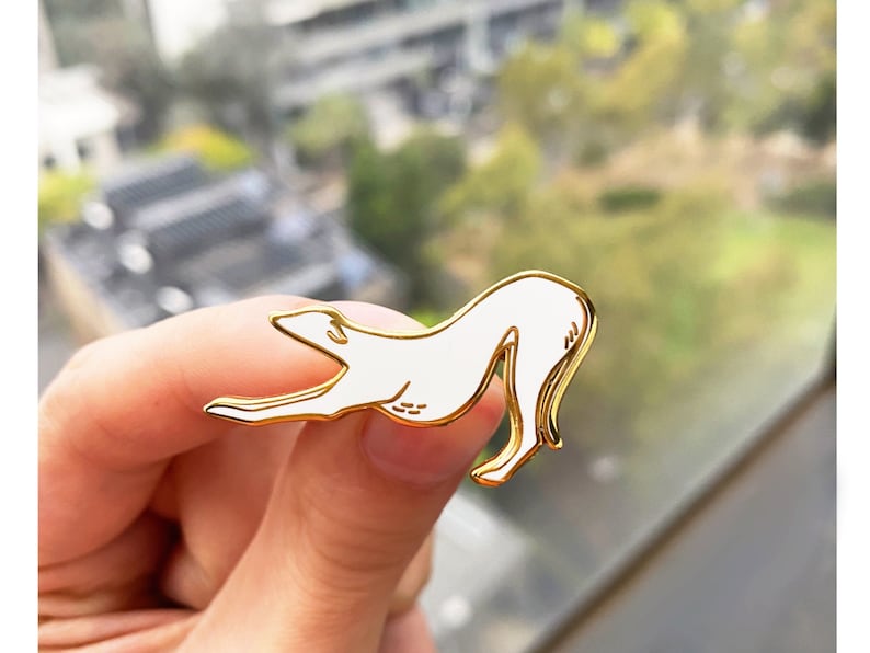 Pode incluir: Um broche de esmalte branco de um galgo em posi&ccedil;&atilde;o de corrida. O broche &eacute; dourado e tem um contorno detalhado.