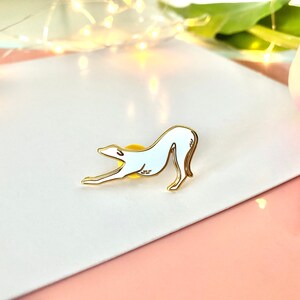 Pode incluir: Um broche de esmalte branco de um c&atilde;o galgo esticando-se, com contornos dourados. O broche est&aacute; sobre uma superf&iacute;cie branca.