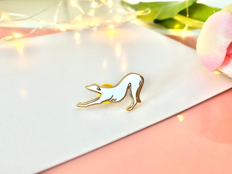 Pode incluir: Um broche de esmalte branco de um c&atilde;o galgo esticando-se, com contornos dourados. O broche est&aacute; sobre uma superf&iacute;cie branca.