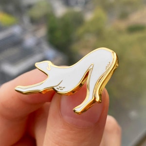 Pode incluir: Pin de esmalte dourado com uma silhueta branca de um c&atilde;o em uma pose brincalhona.