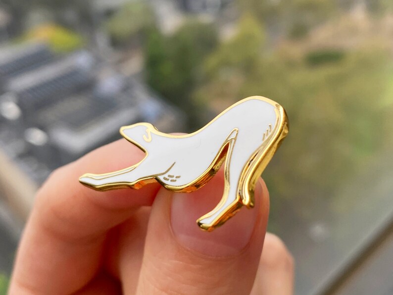 Pode incluir: Pin de esmalte dourado com uma silhueta branca de um c&atilde;o em uma pose brincalhona.