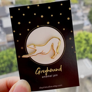 Pode incluir: Pin de esmalte dourado de um galgo em um cart&atilde;o preto com estrelas douradas. O cart&atilde;o diz "Greyhound enamel pin" e "dayzistudios.etsy.com".