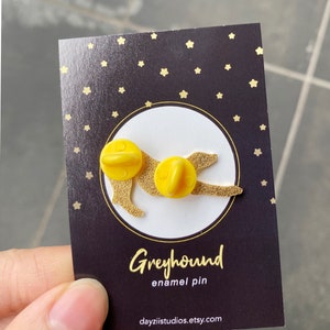 Pode incluir: Pin de esmalte amarelo de um galgo com fundo preto e estrelas douradas. O pin diz "Greyhound enamel pin" e "dayzlistudios.etsy.com".
