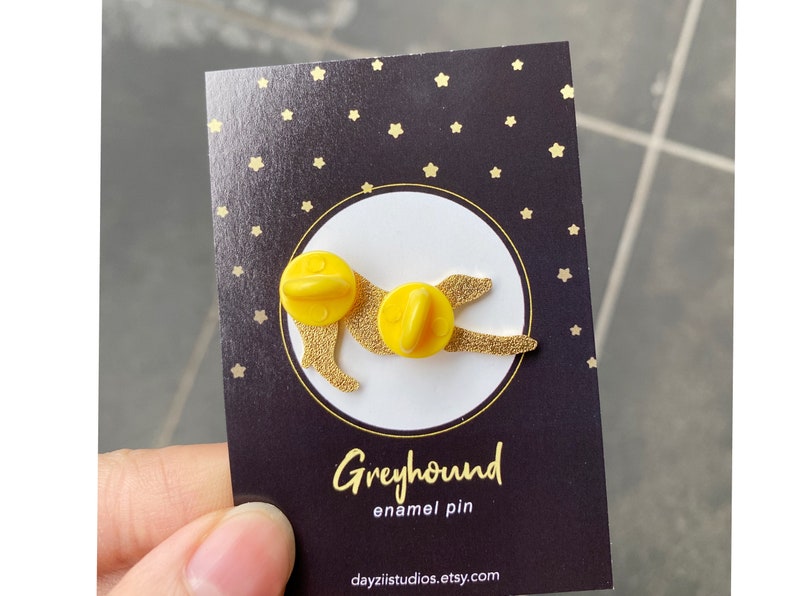 Pode incluir: Pin de esmalte amarelo de um galgo com fundo preto e estrelas douradas. O pin diz "Greyhound enamel pin" e "dayzlistudios.etsy.com".