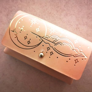 Pode incluir: Uma carteira de couro rosa claro com um design em relevo dourado de uma lua crescente, nuvens e estrelas.