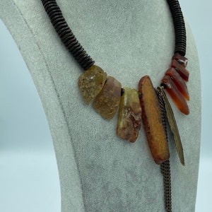 Op de afbeelding: Een statement ketting met een donkerbruine kralen kraag. De ketting heeft een reeks amberkleurige, onregelmatig gevormde stenen en een bronzen veervormige hanger met een kettingkwast. Gepresenteerd op een grijze mannequin.