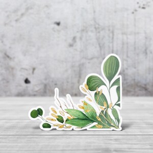 Peut inclure: Un autocollant avec une illustration à l'aquarelle de feuilles vertes et d'accents dorés. L'autocollant a une bordure blanche et est placé sur une surface en bois avec un fond gris. Le design est botanique.