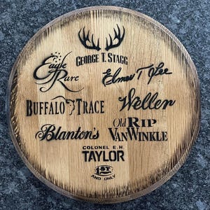May include: A wooden barrel head with a variety of bourbon brands engraved in black lettering, including Eagle Rare, George T. Stagg, Elmer T. Lee, Buffalo Trace, Weller, Blanton's, Old Rip Van Winkle, Colonel E.H. Taylor, and 1st and Only.