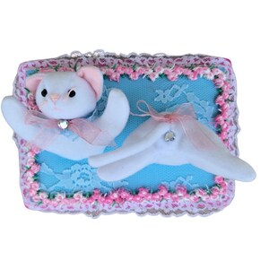 May include: A rectangular decorative plaque featuring a bright blue lace background framed by a pink floral border. Attached to the front are two white fabric cat-like figures, each adorned with a sheer pink ribbon bow and a small clear rhinestone.