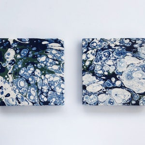 May include: Two square coasters featuring a marbled design in shades of blue, green, and white. The coasters have a unique, abstract pattern.