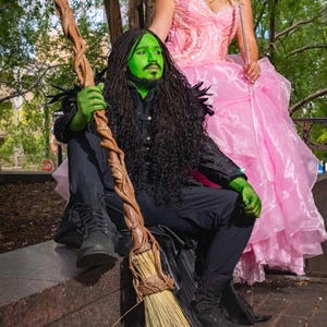 Pode incluir: Uma pessoa com o rosto verde e uma fantasia preta segura uma vassoura marrom. Outra pessoa usa um vestido rosa e segura uma varinha em forma de estrela. A imagem é um cosplay de personagens de uma história de fantasia.