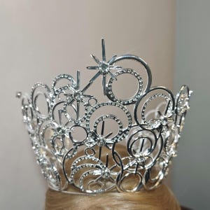 Pode incluir: Uma coroa prateada com um design de estrela. A coroa apresenta múltiplos detalhes de estrelas e círculos, cada um adornado com pequenas pedras preciosas transparentes. A coroa foi projetada para ser usada na cabeça.