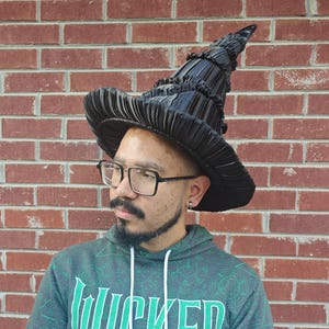May include: A black witch hat featuring a tall, pointed, and pleated cone structure with a wide, ruffled brim. The hat is constructed from a shiny, dark material with distinct vertical folds that create a textured, layered appearance throughout.