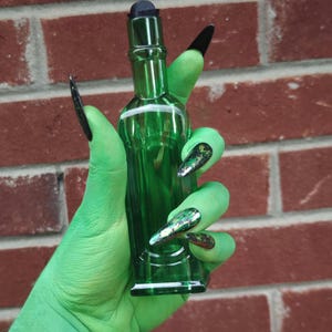 May include: A green glass bottle with a black stopper is held by a green hand with long, black fingernails. The bottle is cylindrical with a slightly flared base. The background is a red brick wall.