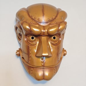 Pode incluir: Uma máscara dourada metálica com um rosto humano estilizado. A máscara apresenta sobrancelhas proeminentes, um nariz grande e aberturas oculares circulares. Rebites e outros detalhes sugerem um design mecânico ou robótico.