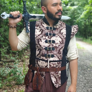 May include: A brown and cream damask patterned vest with black side panels and five front buckle closures. The vest is worn over a cream-colored long-sleeve shirt with rolled sleeves, paired with brown trousers and a brown leather belt with pouches.