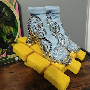 May include: A pair of light blue ankle boots with ornate silver and rhinestone detailing. The heels are gold-colored and the boots rest on a stack of yellow rectangular blocks, reminiscent of a classic movie theme.