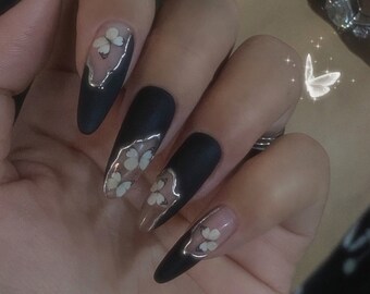 May include: Close-up of a hand with long, black, almond-shaped nails with white butterfly decals. The nails are painted black with a matte finish.