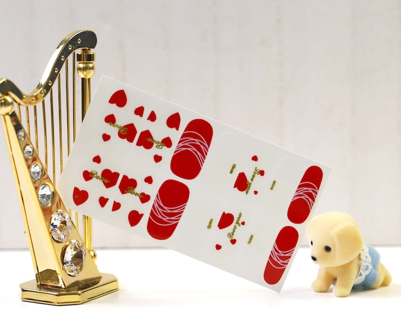 May include: A sheet of red and white nail art stickers with a heart design and the text "Love you". The stickers are on a white background and are next to a gold harp.