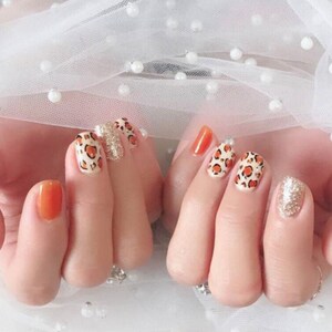 May include: Manicured nails with a variety of designs. Some nails are painted a solid orange, while others feature a leopard print pattern with orange spots on a cream background. One nail has a gold glitter finish.